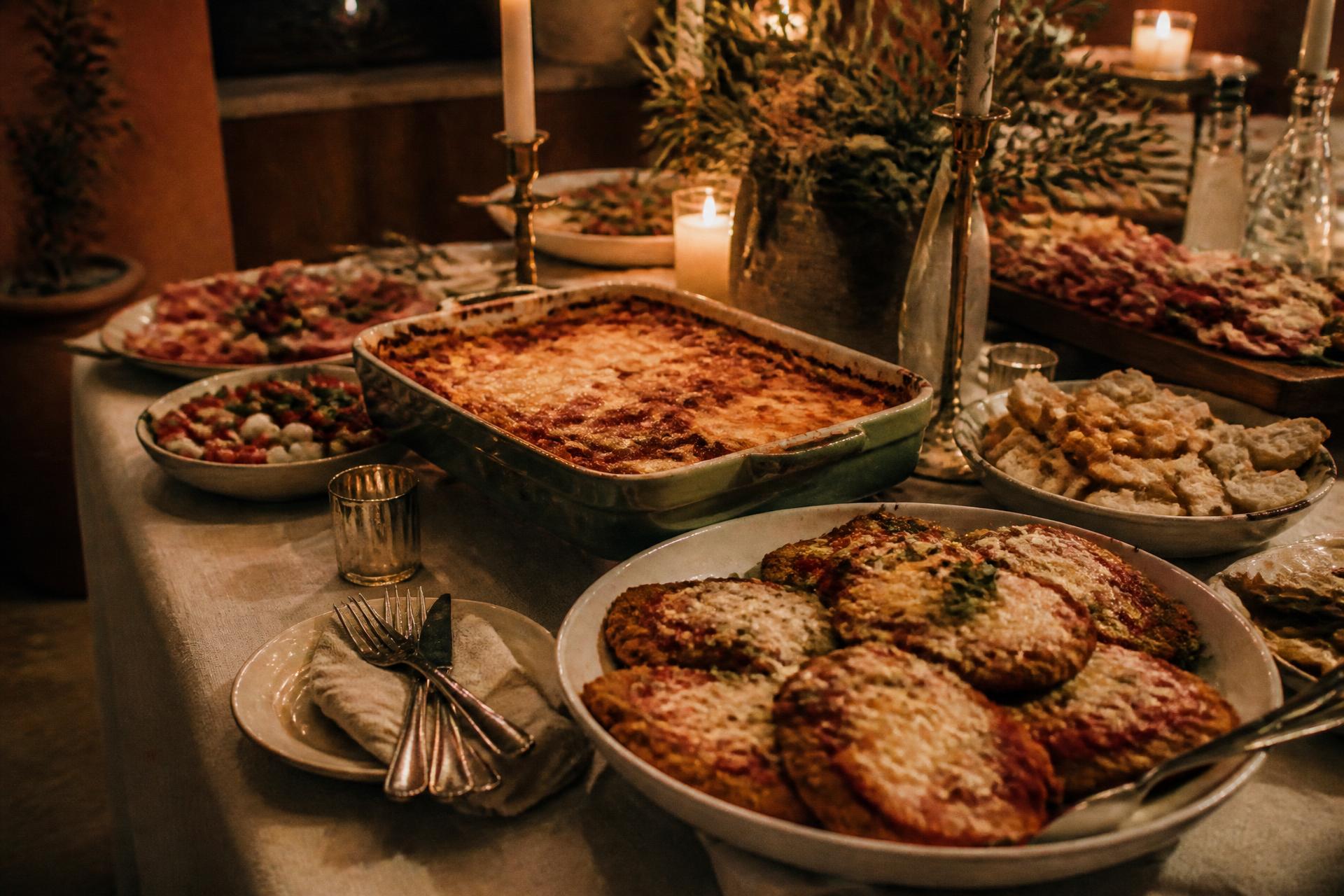 A long table of Italian catering platters — lasagna, chicken parmigiana and antipasti — by candlelight